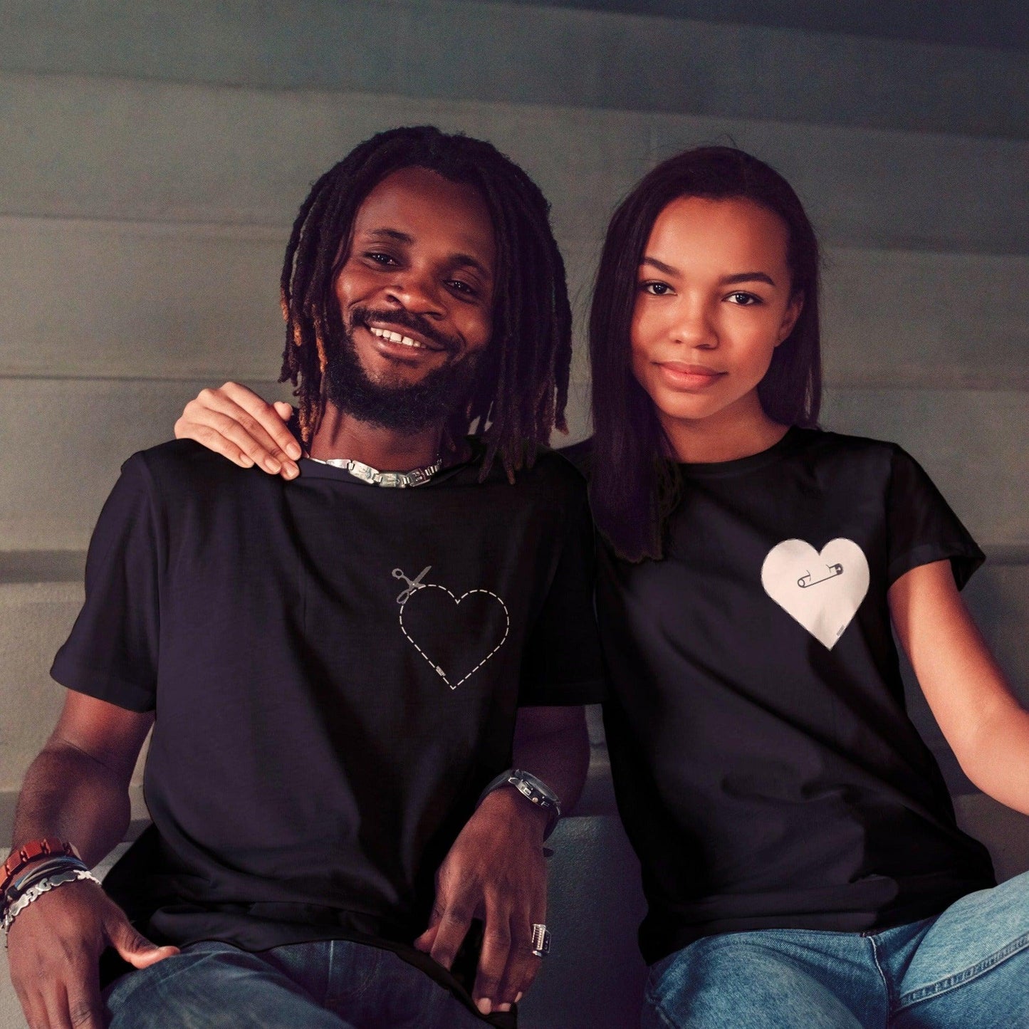 Couple wearing t-shirts. One showing graphic showing how to cut along the dotted outline of a heart with scissors, and the other showing a graphic of a cut out heart pinned with a safety pin