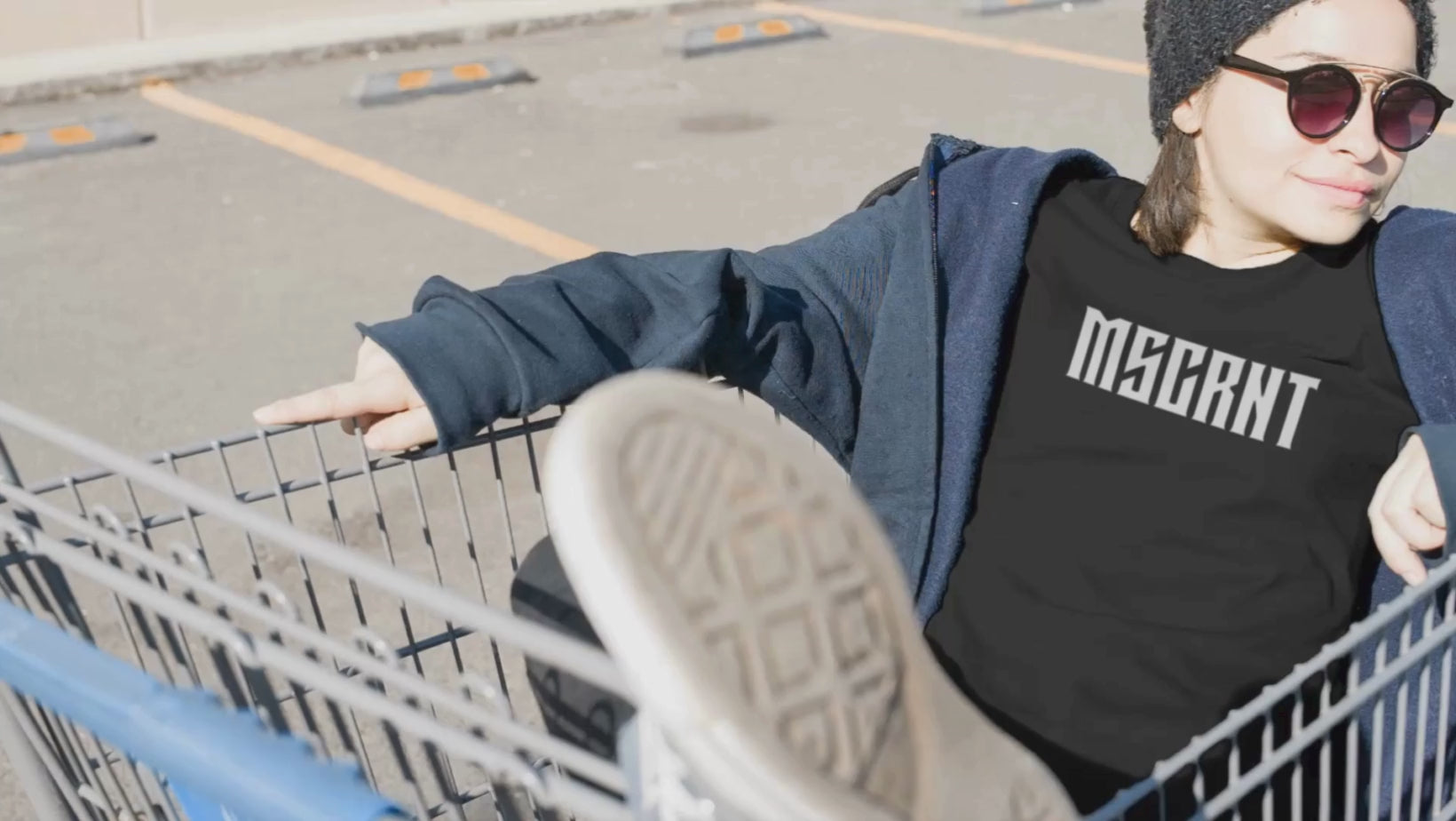 Load video: Girl wearing a MSCRNT black t-shirt, sitting in a shopping trolley that&#39;s moving across the screen.